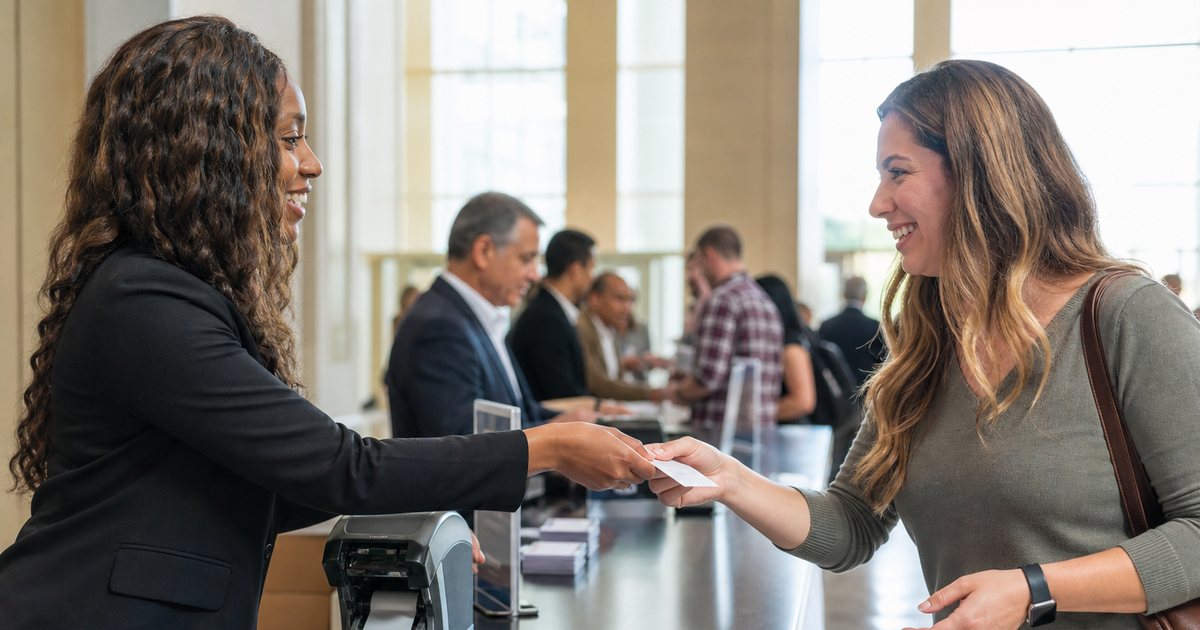 A volunteer at a conference registration desk handing a freshly printed name badge to a smiling attendee, badge printer on the counter, lanyards in a box, morning light through lobby windows