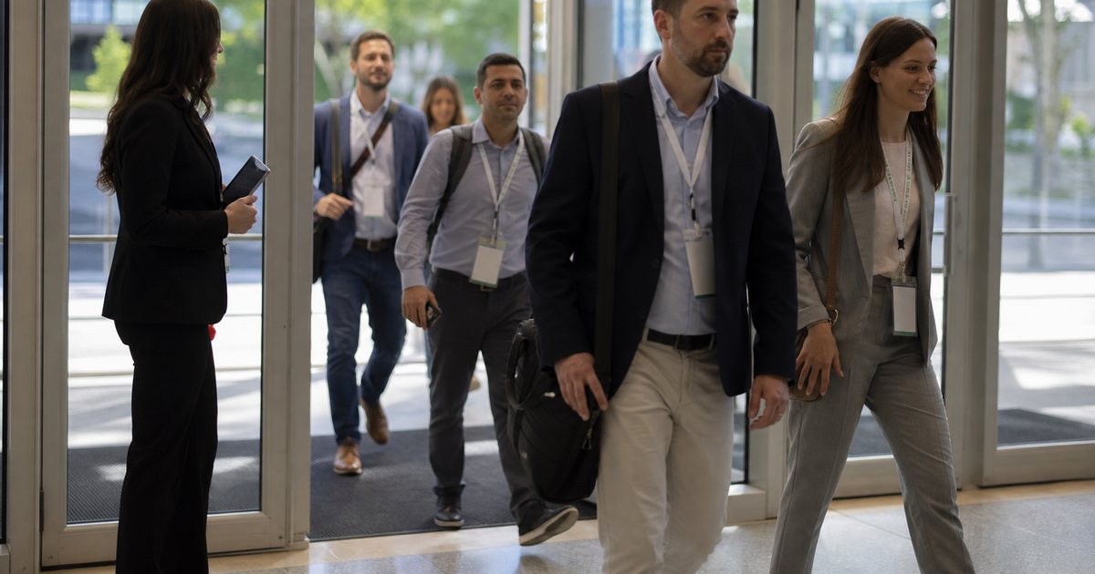 Attendees with lanyard badges walking through the glass entrance of a convention center on a bright morning, a staff member with a handheld device standing beside the doorway