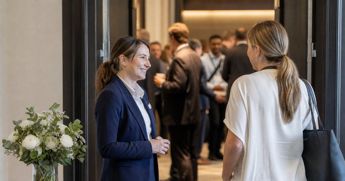 A small side table with fresh flowers and folded lanyards in a quiet hotel foyer, a staff member in a blazer greeting an arriving guest, main conference crowd visible through a doorway